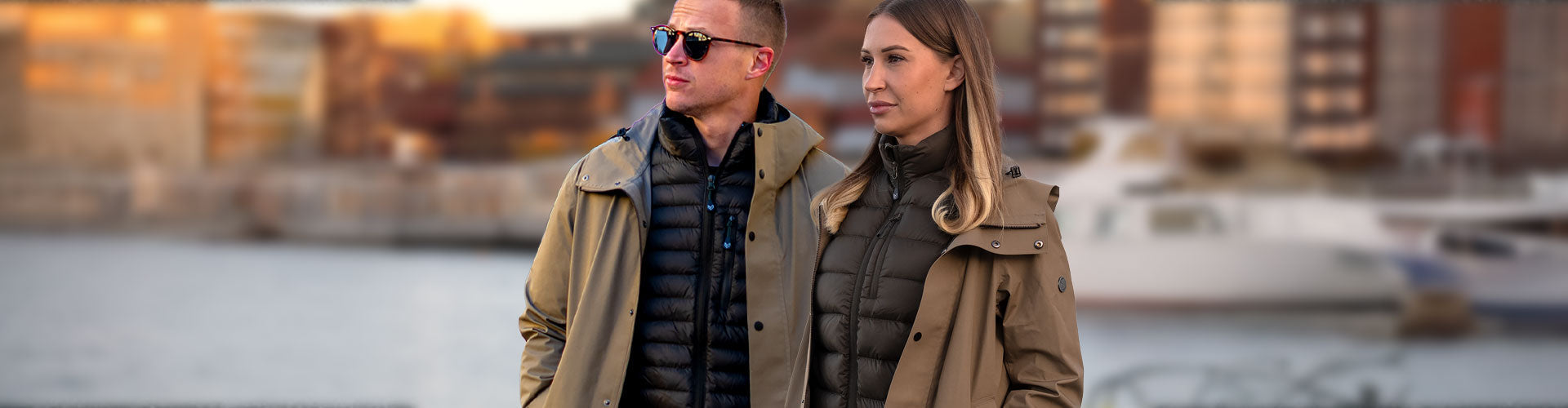 En man och en kvinna iklädda Duster Long Coat från Bright Equipment®. Luleå Norra Hamn i bakgrunden.