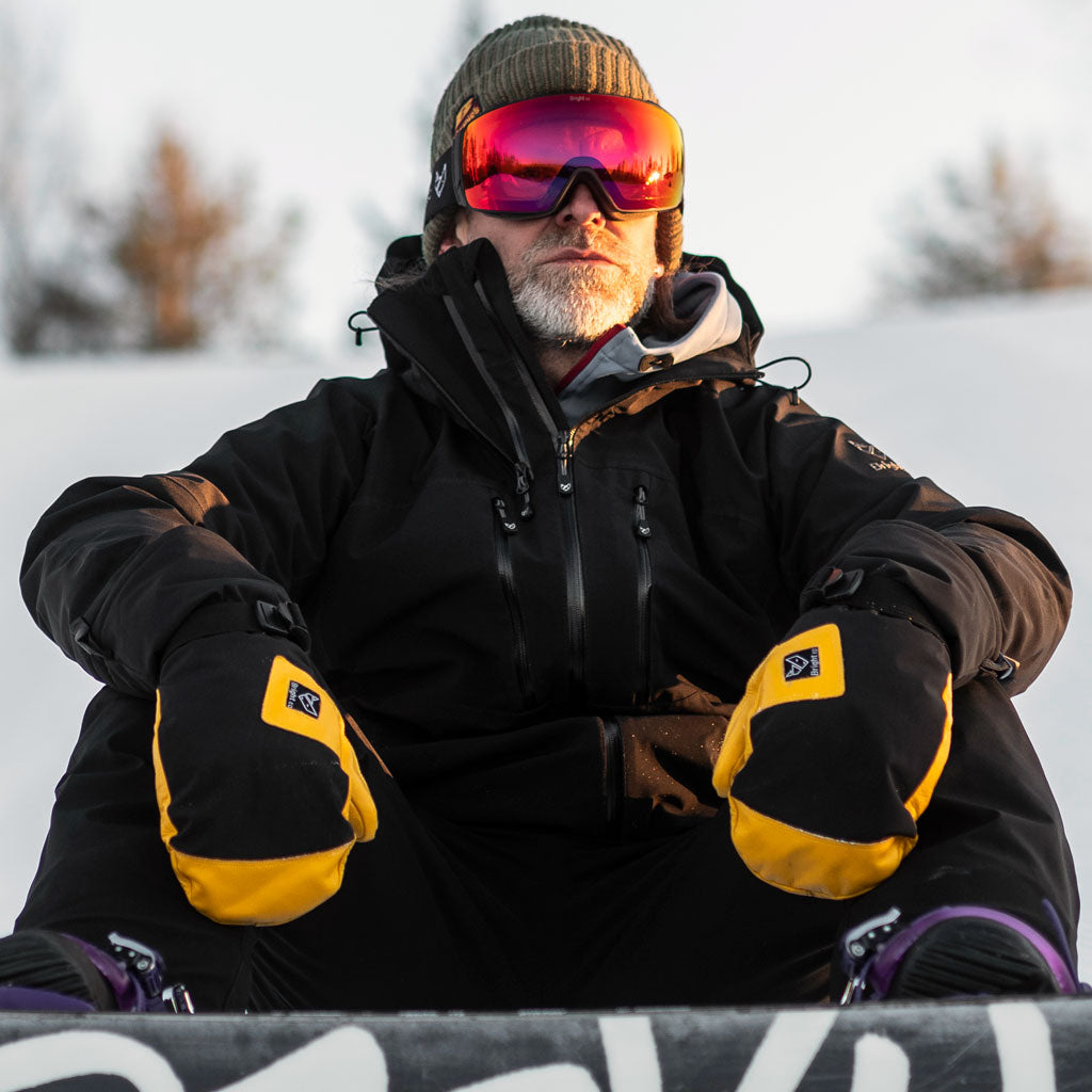 Man med snowboard sitter i skidbacke med handskar, tumhandskar och skaljacka från Bright Equipment.
